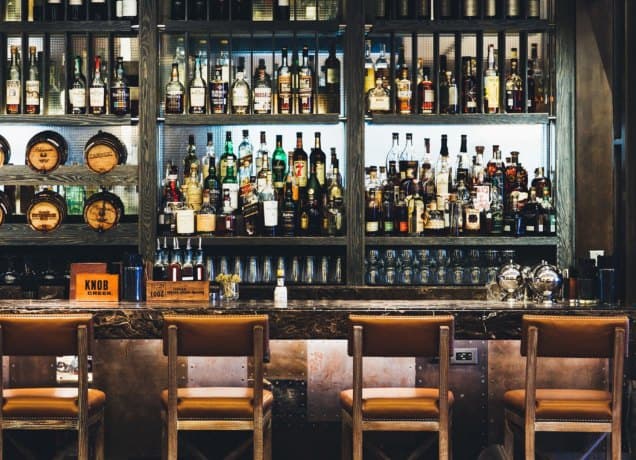loews-philadelphia-review: A photograph of the bar at Bank & Bourbon. It has a large selection of alcohol, a dark countertop and medium stain wooden chairs that face the wall.